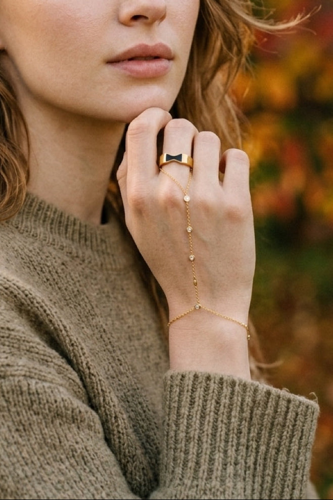 A woman with long light brown hair wearing a textured taupe knit sweater, posing outdoors with a blurred background of autumn foliage. She is wearing a gold ring and a delicate gold chain necklace.