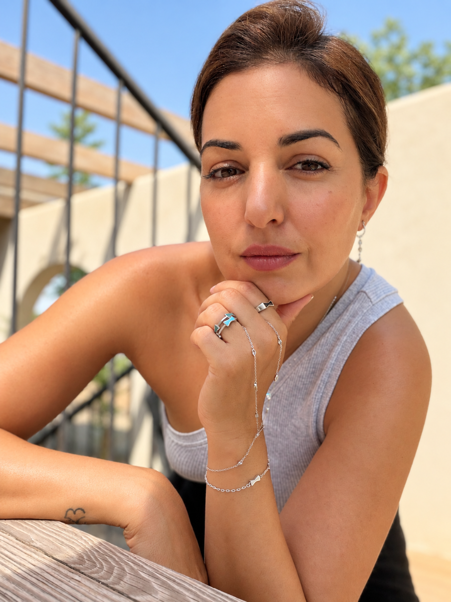 A woman wearing delicate silver chain bracelets and rings, resting her chin on her hand in an outdoor setting with a beige wall and wooden railing in the background.