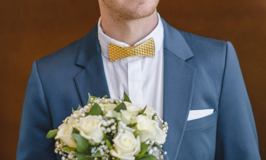 A man in a blue suit, white dress shirt, and yellow patterned bow tie holding a bouquet of white roses, set against a warm brown background.