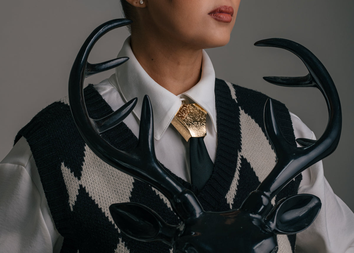 A woman in a black and white argyle sweater vest, white shirt, and black tie holds a glossy black deer head sculpture with antlers against a plain gray studio background.