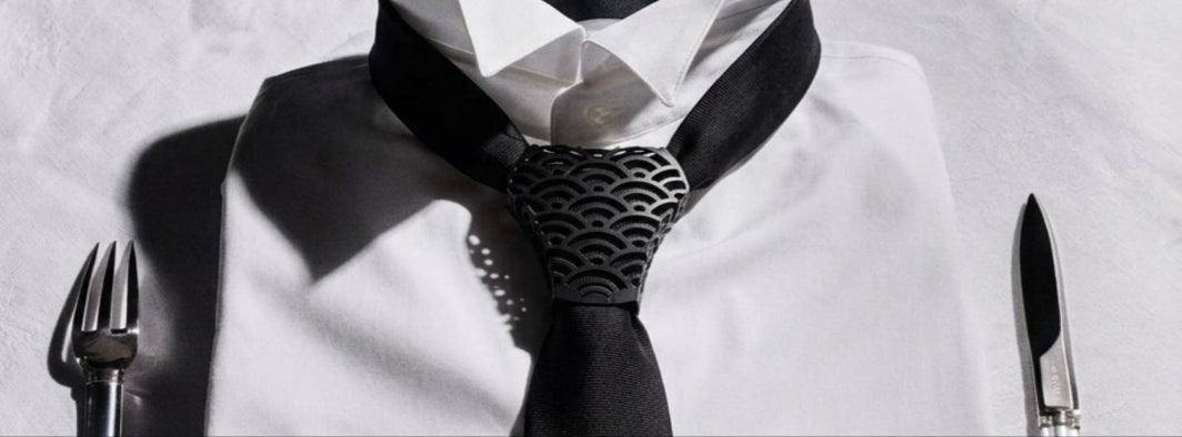 A styled close-up of a white dress shirt collar with a black necktie and a patterned black-and-white tie at the knot, set against a white shirt background with a fork on the left and a knife on the right.