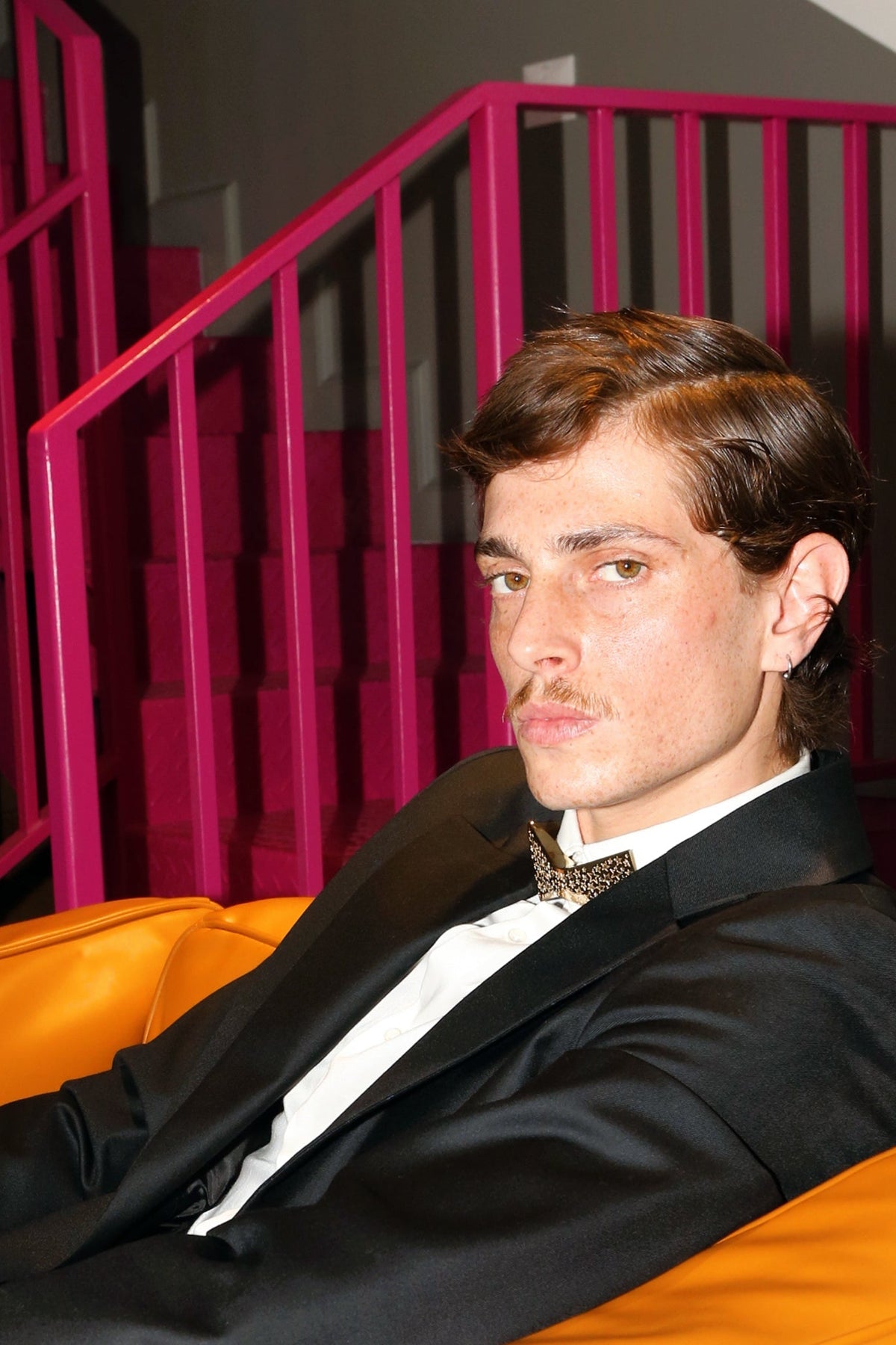Man in a black suit with a bow tie is sitting on an orange couch against a pink staircase.