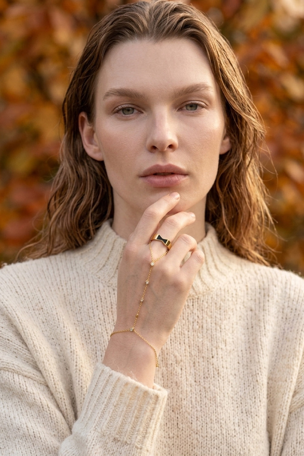 A woman with light brown hair wearing a cream-colored knit sweater, gold chain bracelet, and gold ring, posing in front of a blurred background of autumn leaves.