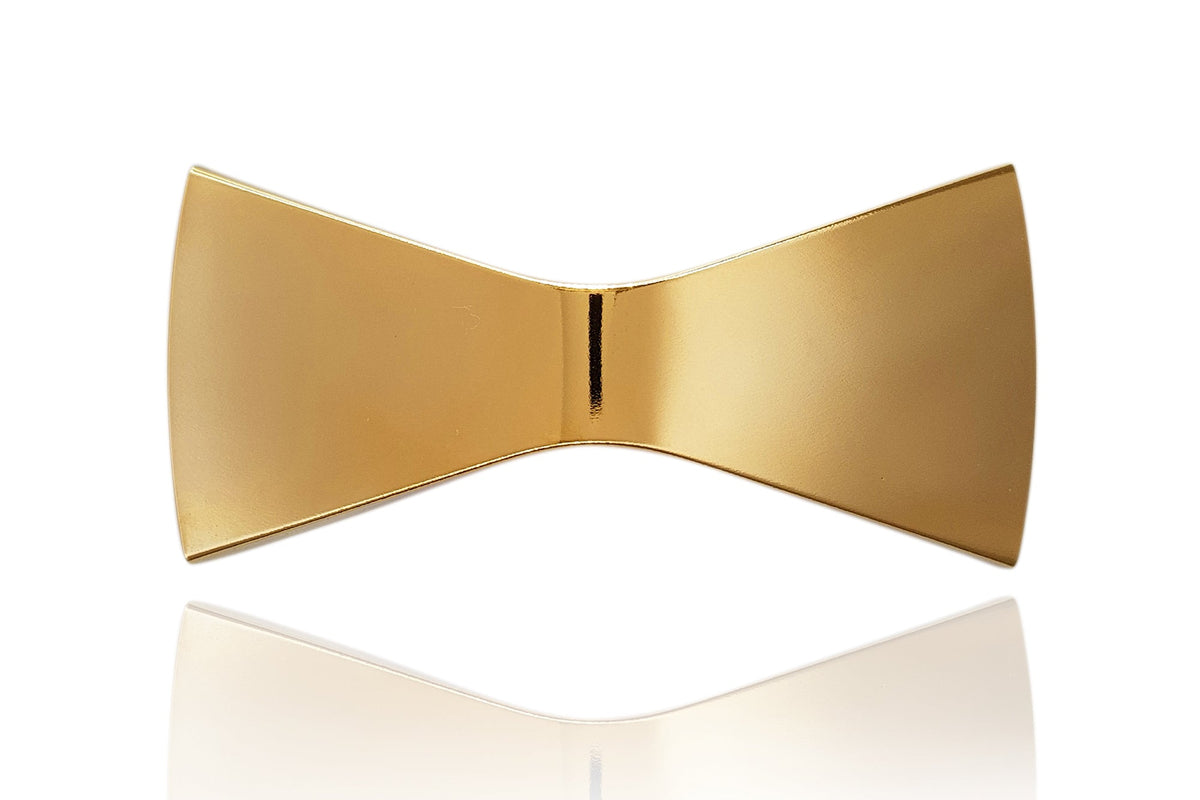 A modern, sculptural gold bow tie-shaped accessory with a polished metallic finish, displayed on a clean white background with a soft reflection below.