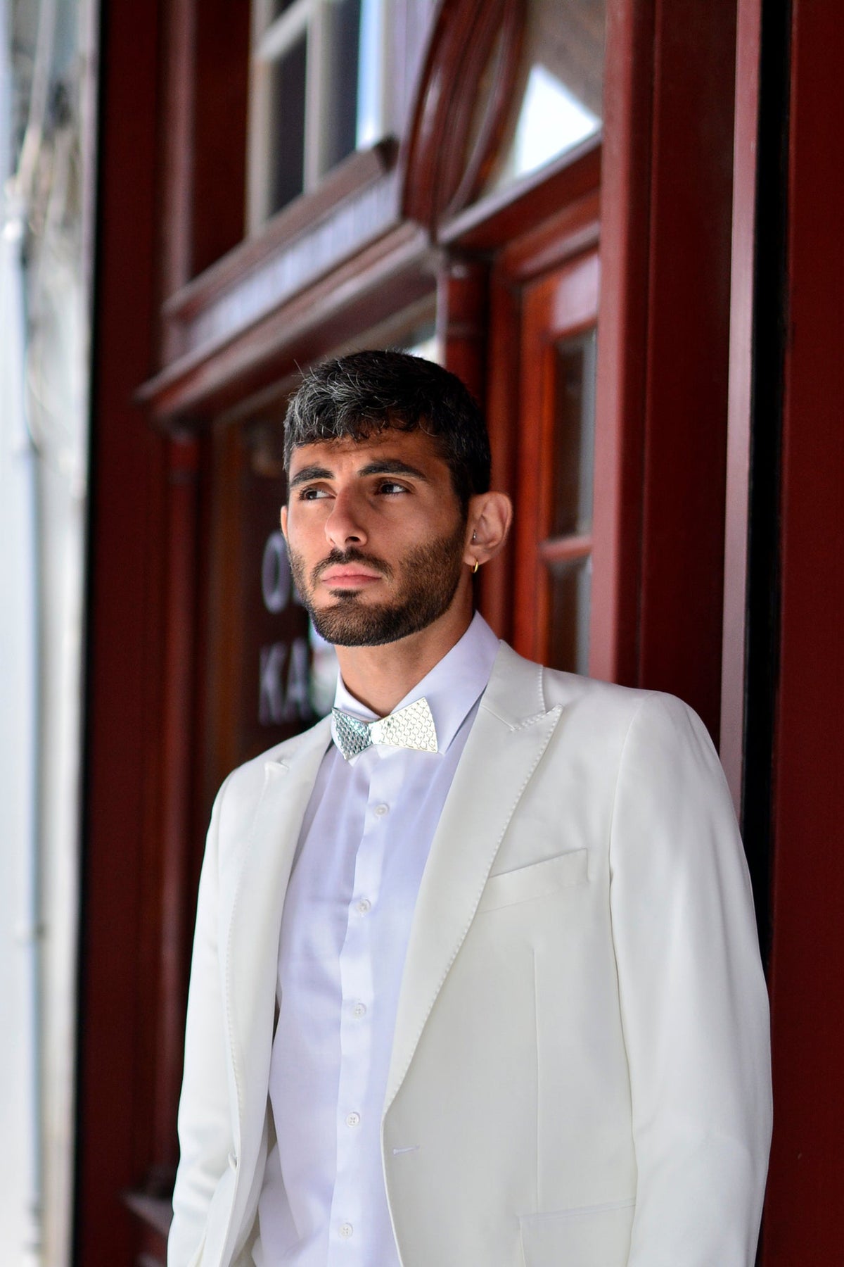A man in a White suit and silver bow tie against a red building.
