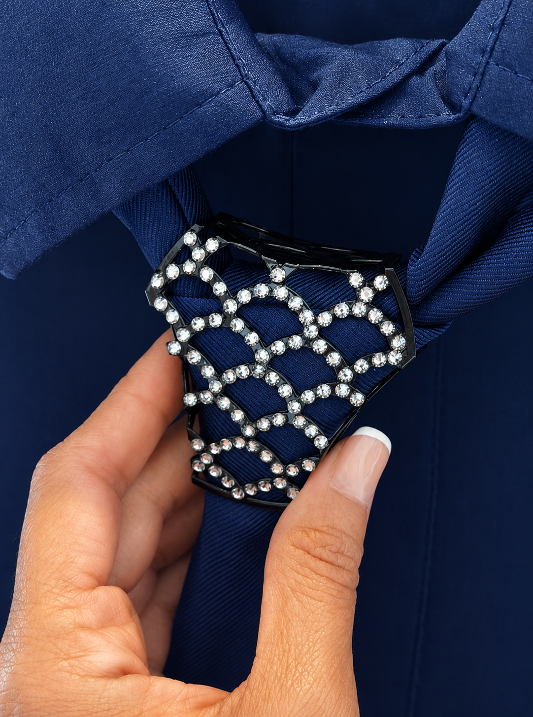A close-up of a hand adjusting a navy blue necktie with a black bow tie clip decorated with a silver rhinestone lattice pattern.