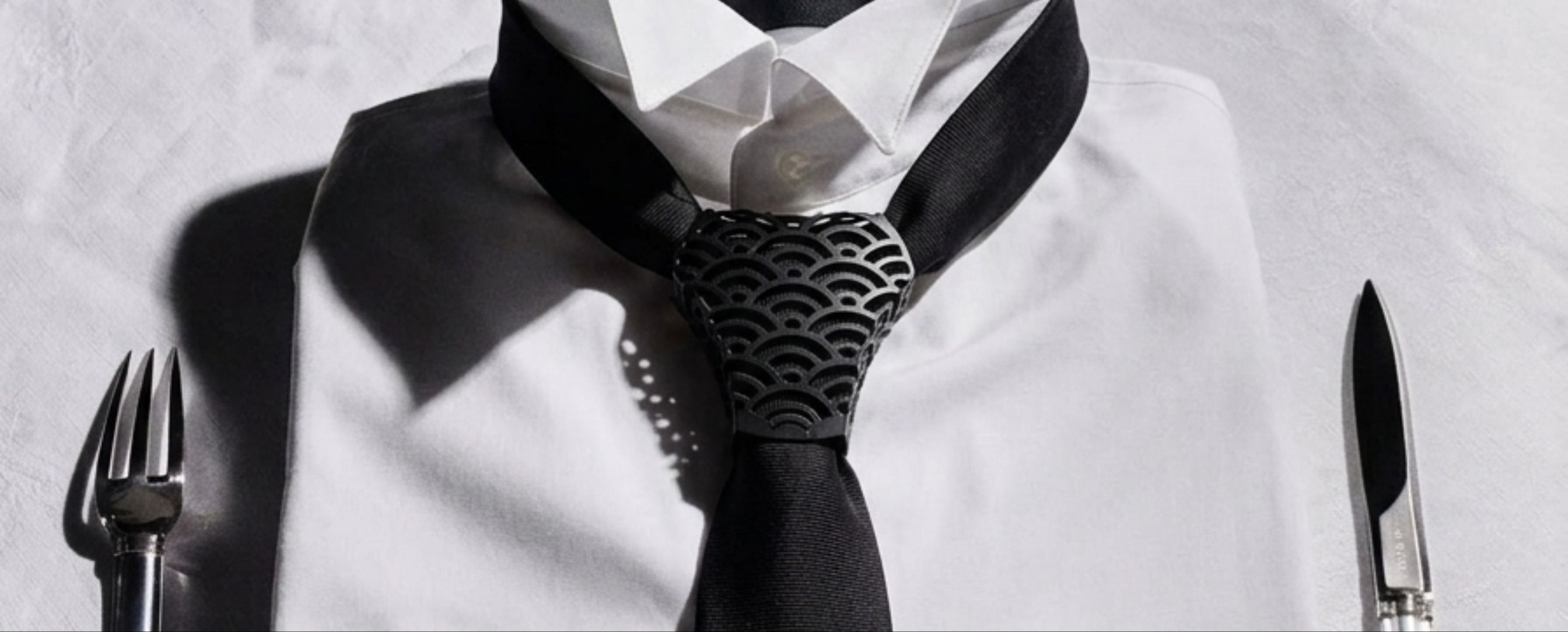 A styled close-up of a white dress shirt collar with a black necktie and a patterned black-and-white tie at the knot, set against a white shirt background with a fork on the left and a knife on the right.