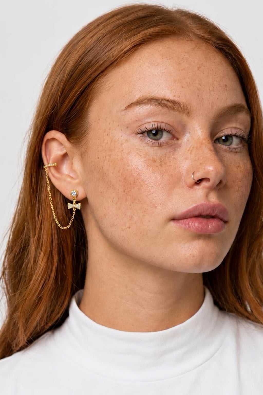 A close-up of a woman with red hair wearing a delicate gold ear cuff with a small butterfly charm and a thin gold chain, paired with a white mock neck top against a light gray background.