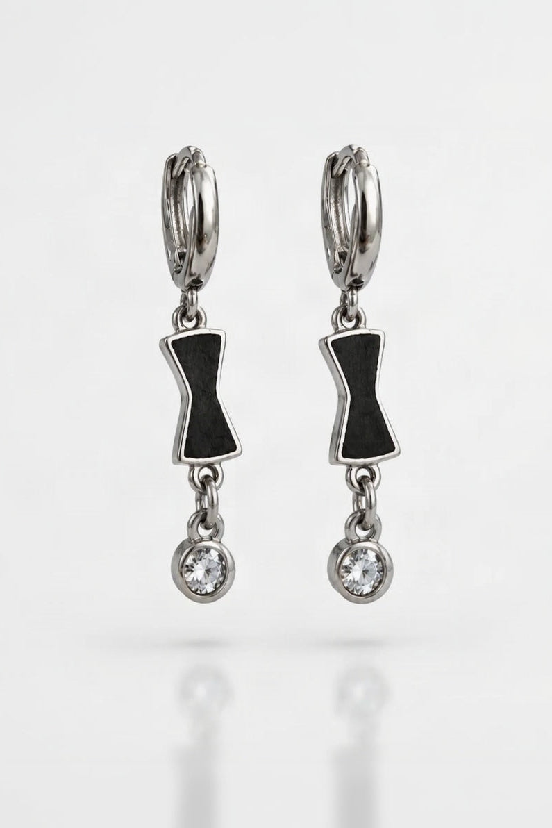 A pair of silver huggie hoop earrings with black bow tie-shaped charms and a dangling clear crystal, shown on a white reflective background.