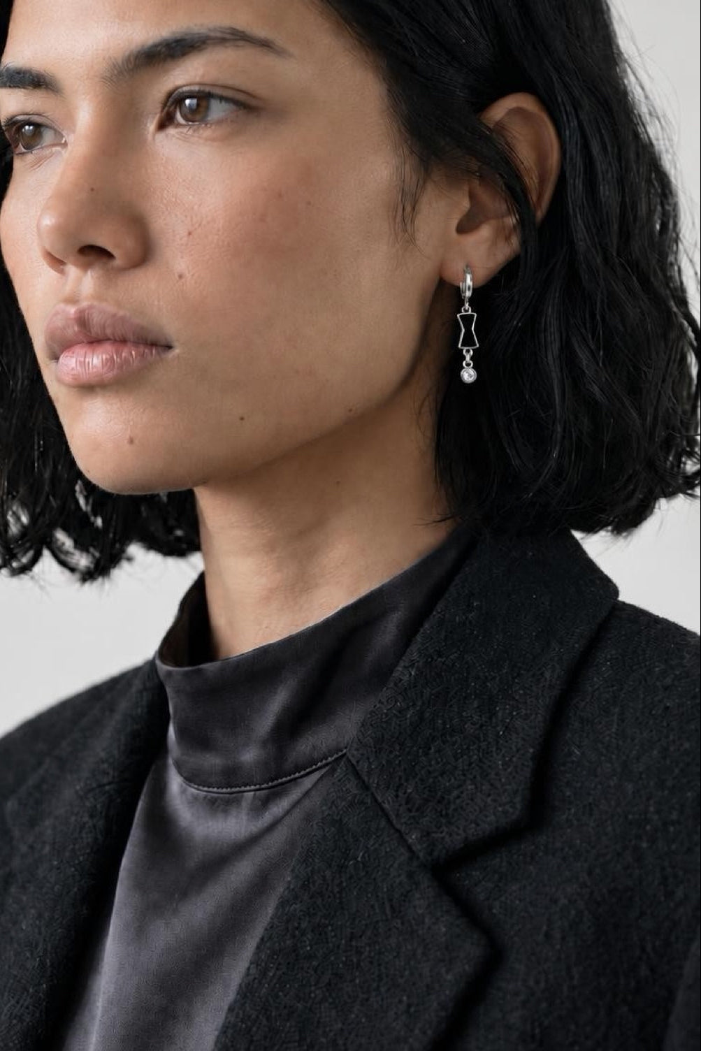 A close-up portrait of a woman with short dark hair wearing a delicate silver dangle earring with a small clear stone, paired with a black satin top and dark blazer against a light gray background.