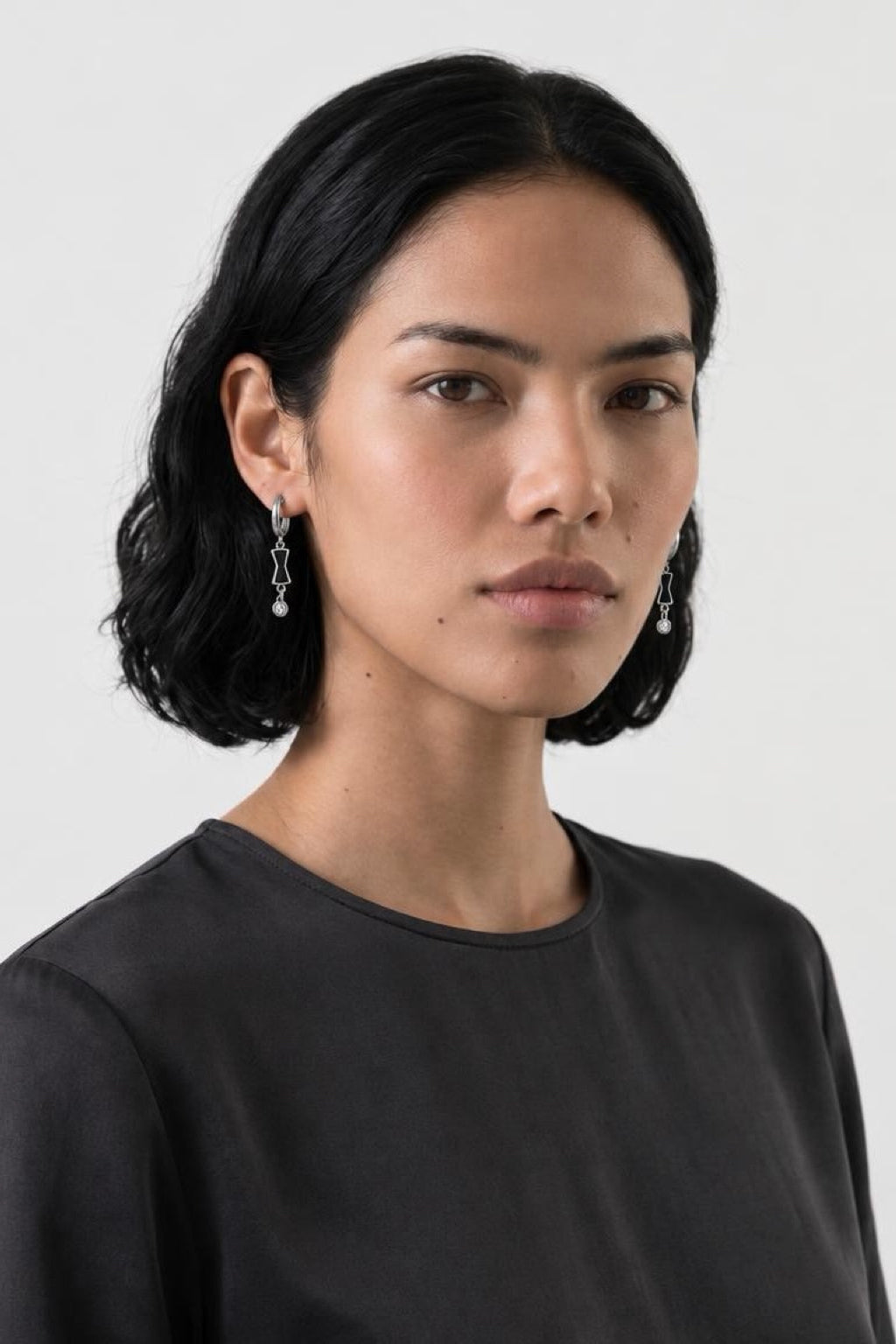 A woman with short dark hair wearing a dark charcoal gray crewneck top and delicate silver drop earrings, photographed against a light gray studio background.