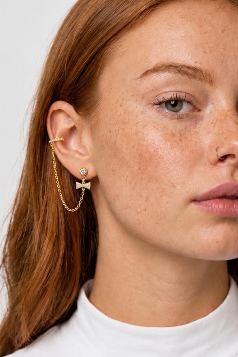 A close-up of a woman's ear wearing a delicate gold ear cuff with a chain drop and a small bow-shaped stud earring with a clear stone.
