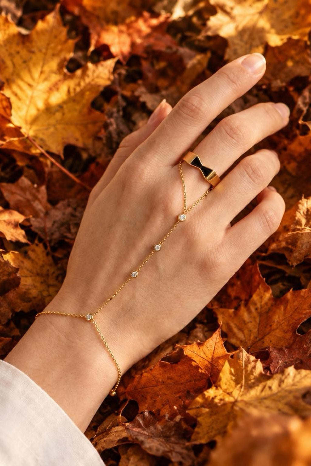 A gold chain hand bracelet and matching ring worn on a hand resting on autumn leaves. The hand bracelet features a delicate chain draped across the fingers with small white pearl-like beads, and the ring has a geometric gold design.