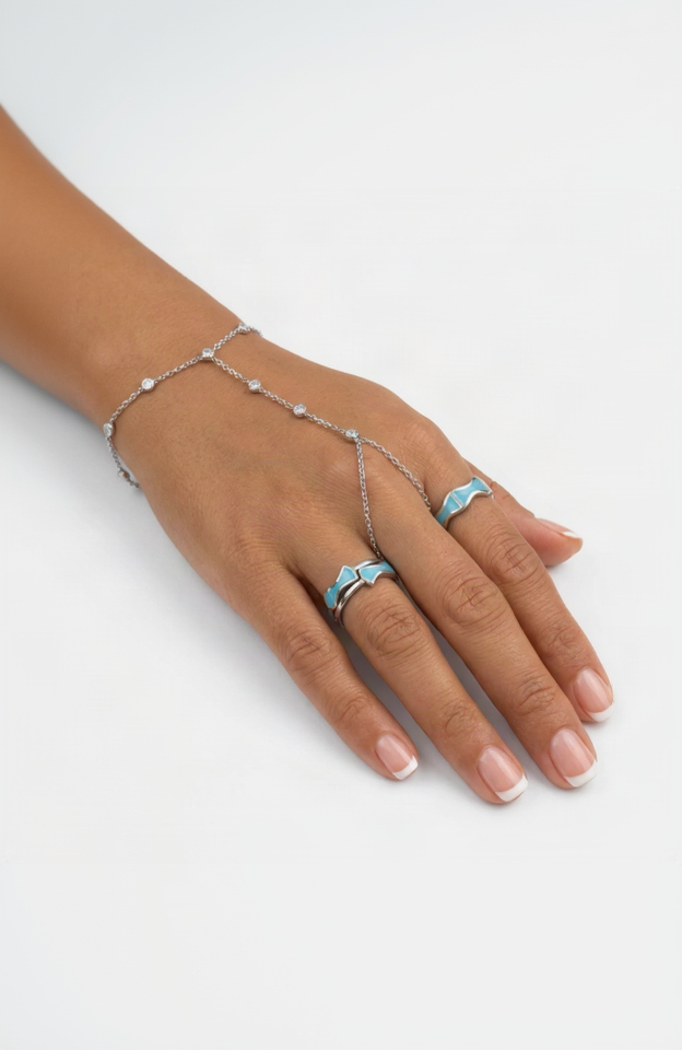 A hand wearing a delicate silver chain hand bracelet with small clear stones and matching silver rings with light blue inlay, shown against a clean white background.