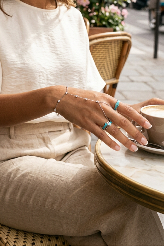 A hand wearing a delicate silver chain hand bracelet with small clear stones and matching silver rings with light blue inlay, shown against a clean white background.